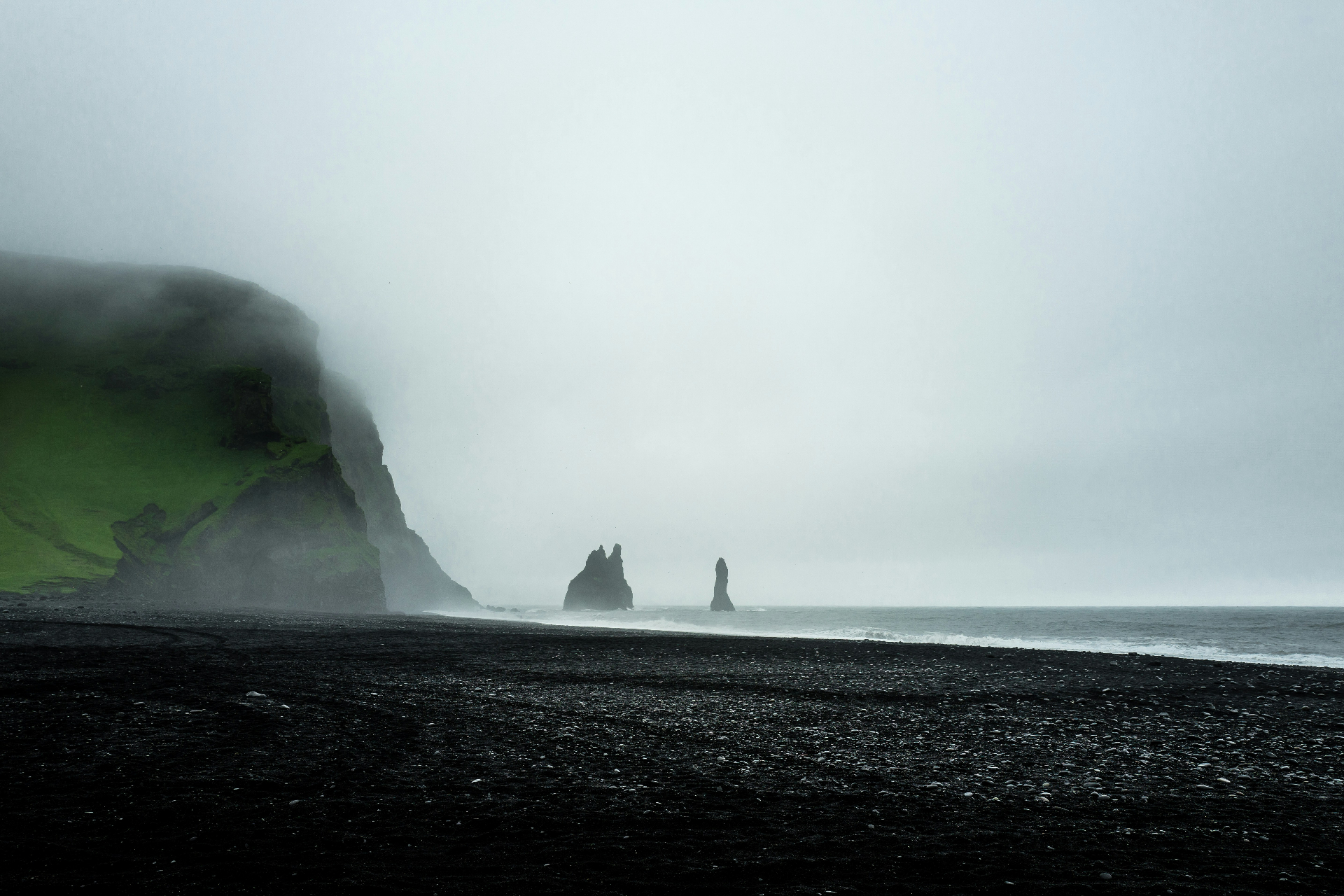 A misty black-sand beach opens toward towering sea stacks in the North Atlantic: the fog and the shore, a beginning.